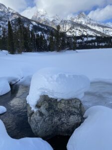 Snow covered lake and boulder with Teton Mountains behind.