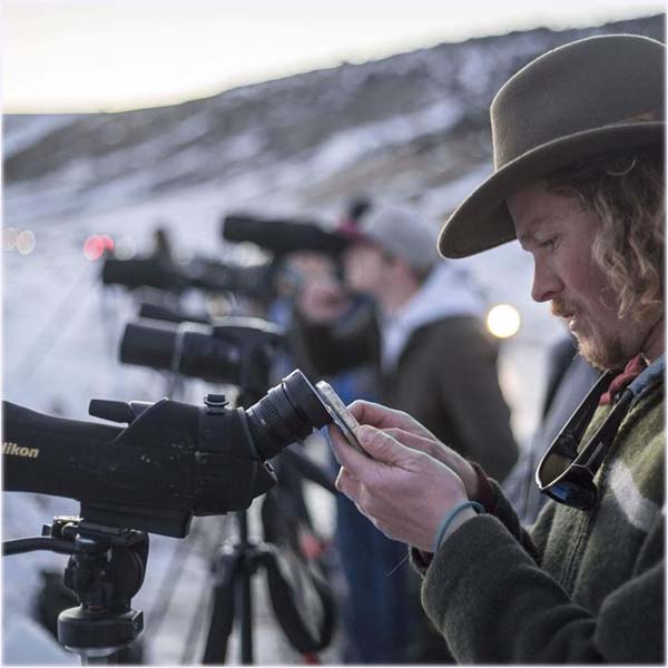 Trevor Bloom looks through a spotting scope with crowd behing him.